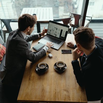 Two businessmen talking about new opportunities sitting with laptop at desk, planning project, considering business offer, sharing ideas while drinking coffee together